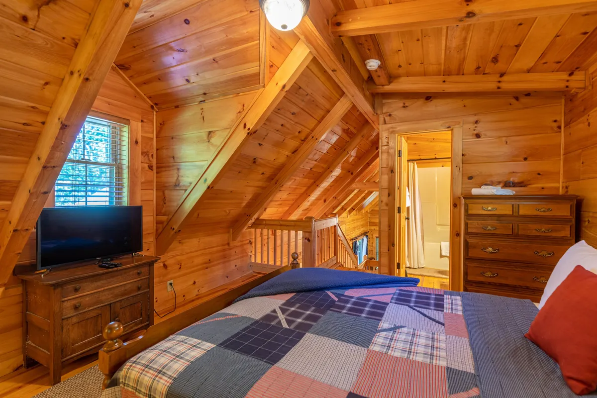 A spacious loft area features a bed with a colorful patchwork blanket, television, wooden dresser, and overhead beams. This inviting space is perfect for relaxation, watching TV, or getting cozy after a day outdoors.
