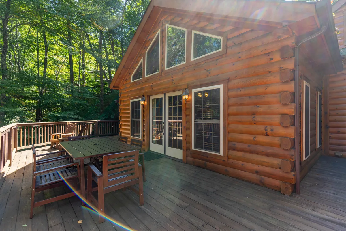 A broad view of the log cabin's exterior shows a large deck outfitted with a dining set. The deck is surrounded by lush trees, and large windows in the cabin bring in natural light. The outdoor dining area is ready for alfresco meals, and the overall mood is tranquil and welcoming, inviting social gatherings or peaceful breakfasts in the fresh mountain air.