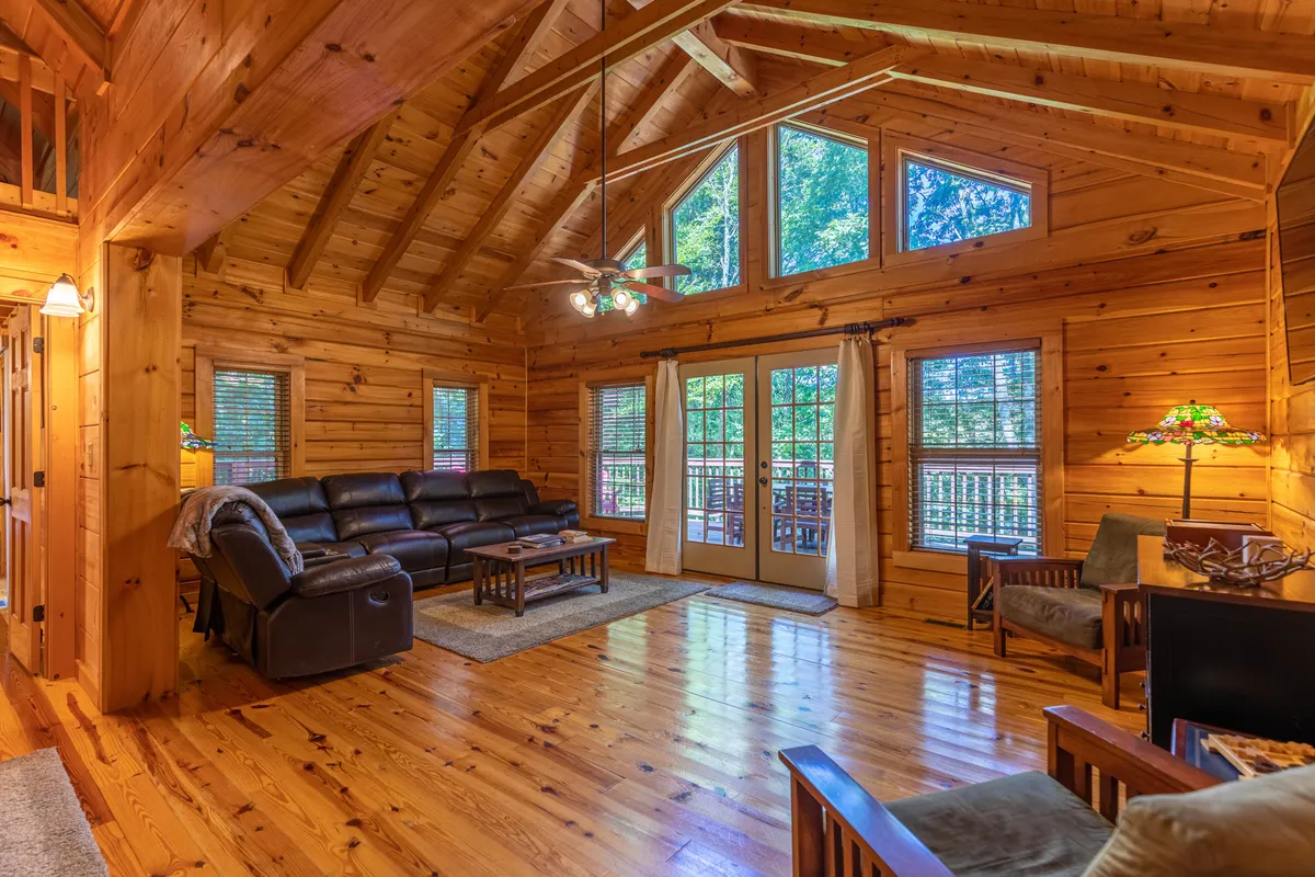 This image captures a cozy and bright living area with exposed wooden beams, large windows that reveal scenic outdoor views, and comfortable seating arranged around a coffee table. Sunlight streams through the windows, creating a seamless connection between the indoors and the surrounding landscape.