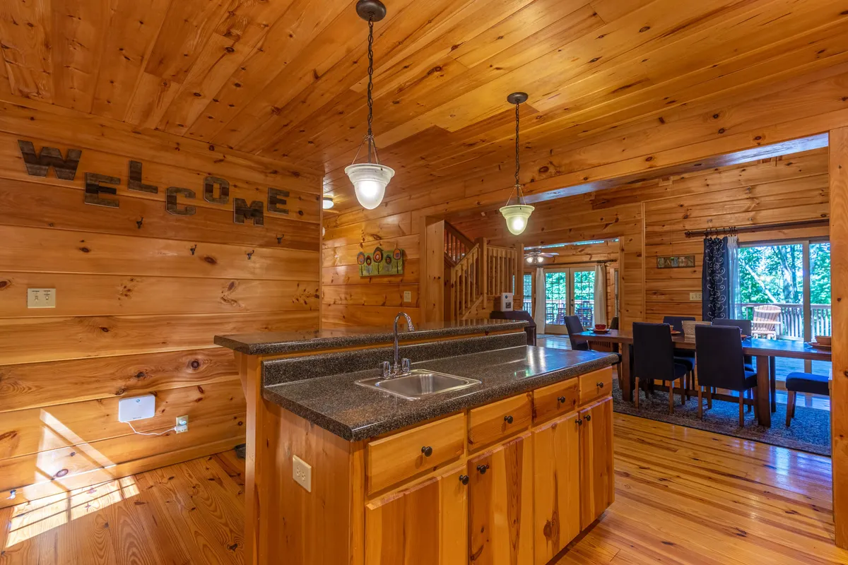 This image centers on the kitchen island with a sink, showcasing a welcoming 'WELCOME' sign and a view of the dining area and wooden staircase. The space is bright, airy, and well-equipped—ideal for group cooking and casual gatherings.