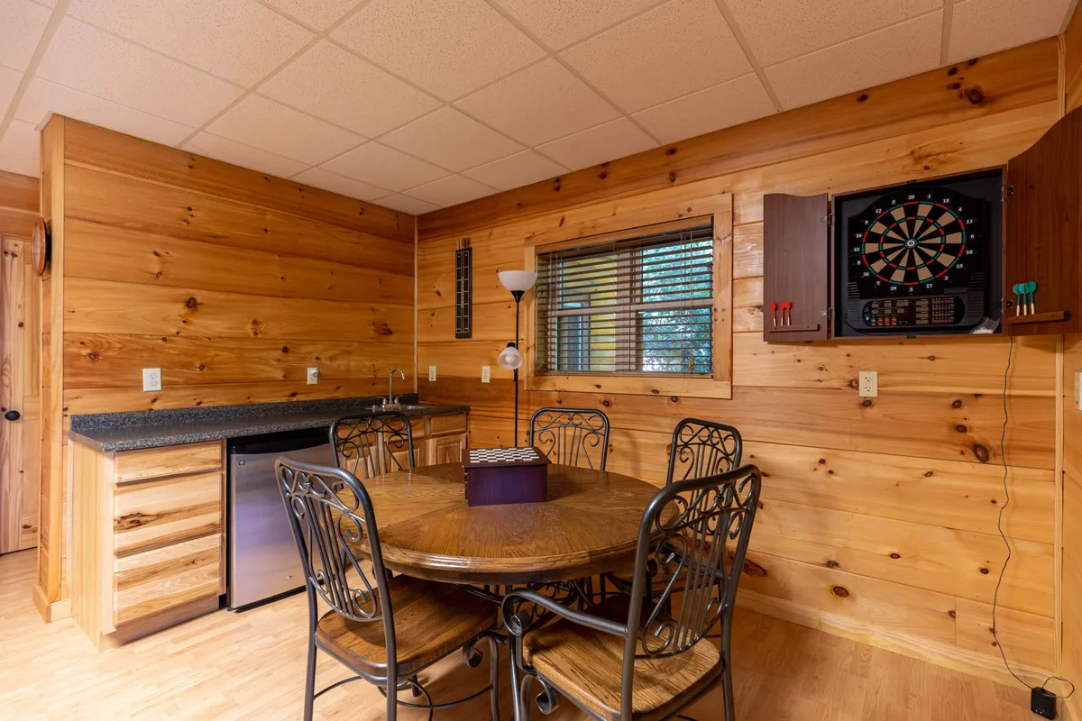 A wooden dining table set near a dartboard and a kitchen counter, framed by rustic wooden walls. The scene is ideal for casual meals, board games, or a friendly round of darts.