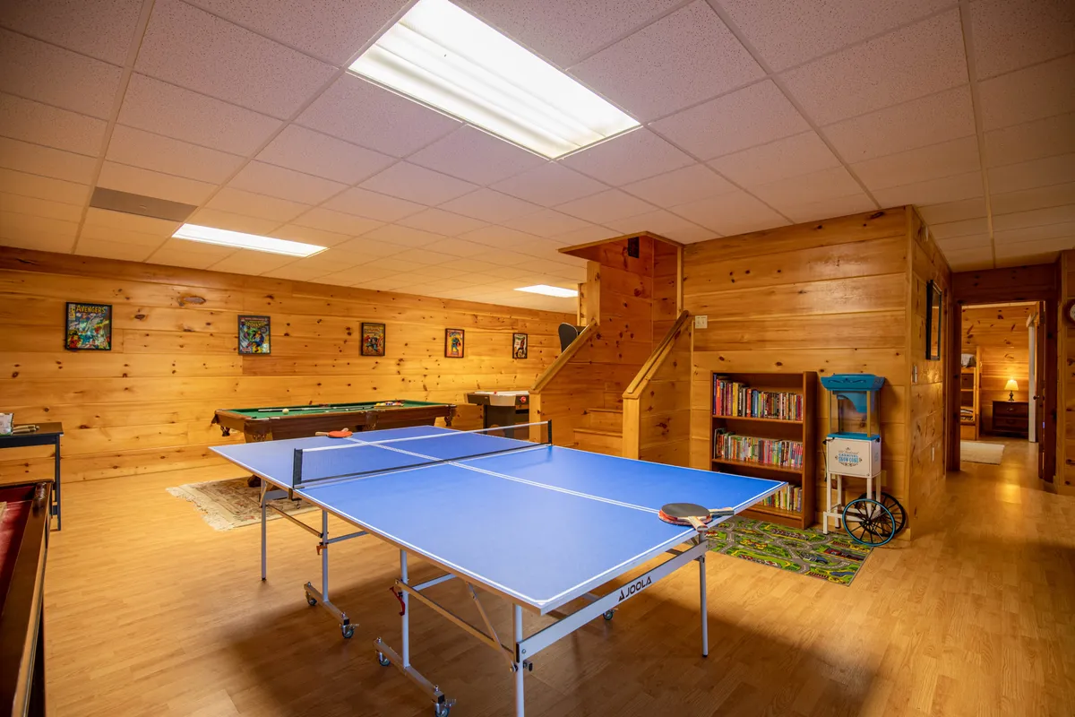 This image captures a spacious game room featuring both a ping pong table and a pool table. Warm wooden paneling and a bookshelf add to the cozy, inviting vibe, while a staircase hints at adjacent living areas. The setup encourages playful competition and socializing, perfectly illustrating the entertainment options available indoors.