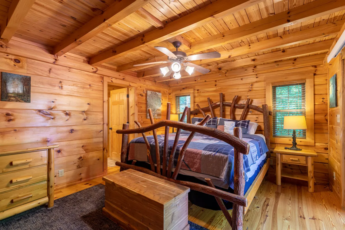 A rustic wooden bed takes center stage in this cozy bedroom, complemented by a nightstand with a soft-glowing lamp, tasteful artwork on the walls, and a wooden dresser. The room exudes warmth, inviting relaxation and restful sleep amid natural textures.