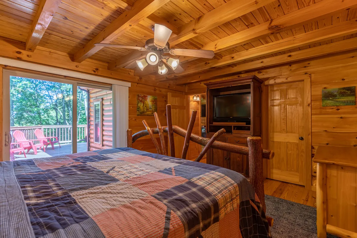 A cozy bedroom with a quilted bed positioned near a sliding glass door, which leads to a wooden deck. The space features exposed wooden ceiling beams and offers tranquil views of the outdoors, inviting natural light into the room.