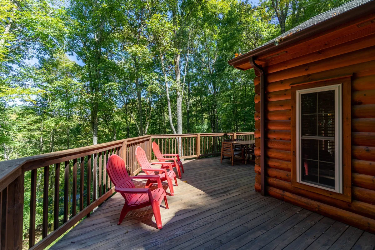 This image features a wooden deck attached to the log cabin, with red Adirondack chairs arranged to face the surrounding trees. The deck is nestled in a peaceful mountain setting, and the inviting chairs suggest a comfortable space to relax, enjoy morning coffee, or watch sunsets. The surrounding greenery and log wall evoke a cozy, secluded feel, and the setup is perfect for gatherings or quiet moments with loved ones and pets.