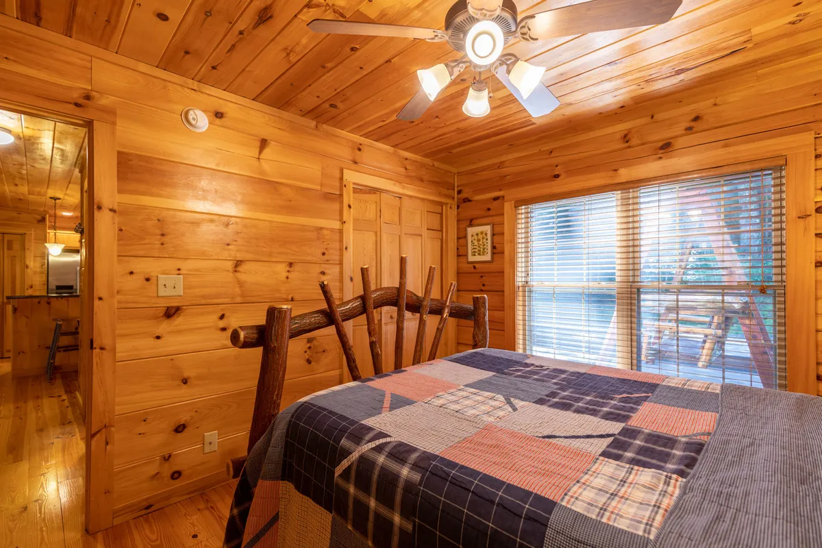 A serene bedroom featuring a wooden bed frame, plaid bedspread, window with blinds, wood paneling, and a ceiling fan. The space feels private and inviting, offering a quiet retreat for relaxation and restful sleep.