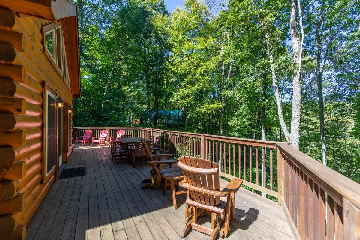This image displays the inviting wooden deck and log cabin facade of Wolf Den Lodge, nestled among tall trees. The spacious deck features comfortable seating areas, perfect for relaxing outdoors and soaking in the peaceful natural surroundings.