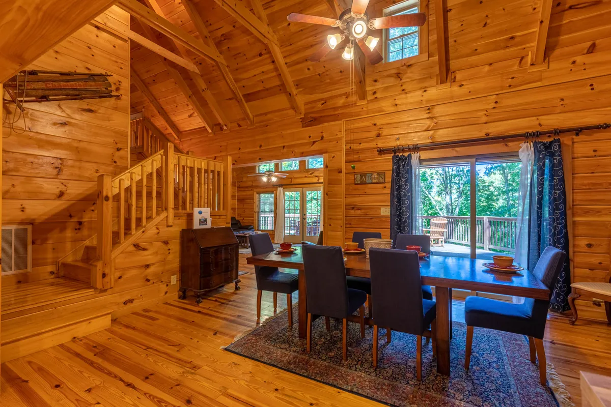 This image features a cozy dining table set near large windows, with exposed wooden beams and a staircase visible. The windows suggest a strong connection to the outdoors and natural surroundings, allowing guests to enjoy views of the changing seasons from the heart of the home. The overall mood is warm and inviting, perfect for gathering after a day of adventure.