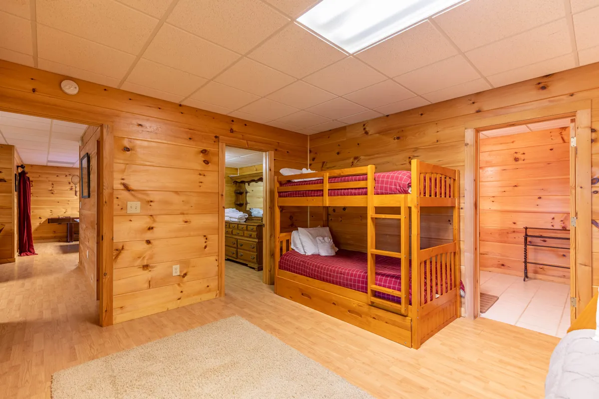 A cozy bunk room with wooden walls, multiple bunk beds, a soft rug, and doorways leading to other areas, creating a friendly and relaxed atmosphere ideal for kids or groups. The arrangement suggests opportunities for memorable sleepovers and group bonding.