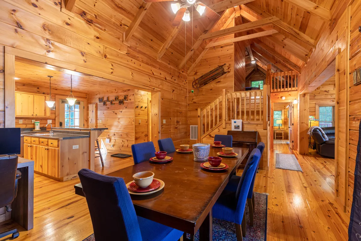 A warm, sunlit dining area with a table set for a meal, adjacent to a wooden staircase and kitchen area. The arrangement invites communal gatherings, family meals, or casual group activities.