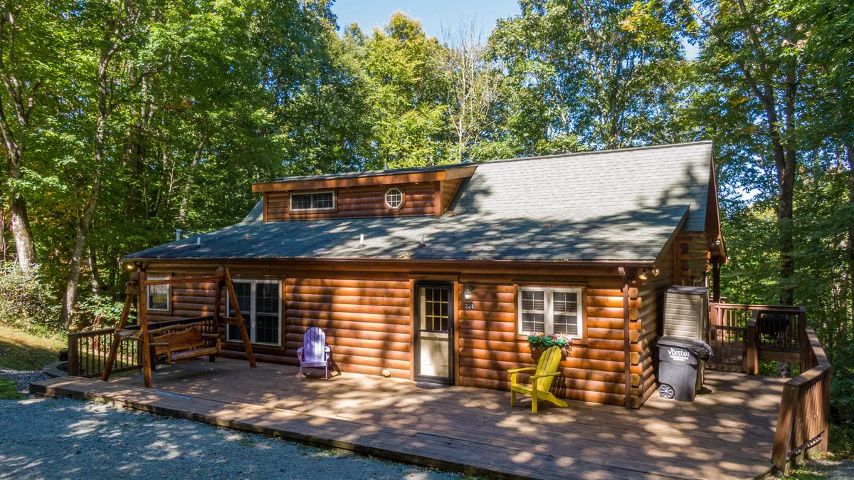 The image showcases a charming log cabin nestled among the trees, featuring a spacious wooden deck adorned with colorful chairs and a swinging bench. The setting captures the warm glow of sunset, inviting guests to relax, gather, and soak in the surrounding mountain scenery.