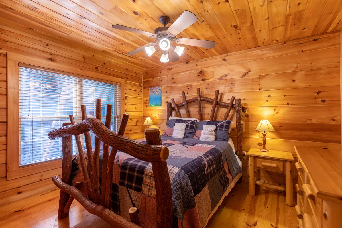 A log bed with plaid bedding is flanked by wooden furniture and a nightstand with a lamp, all set beneath a ceiling fan. The space is brightened by natural light, creating a warm and inviting atmosphere ideal for restful sleep or curling up with a book.